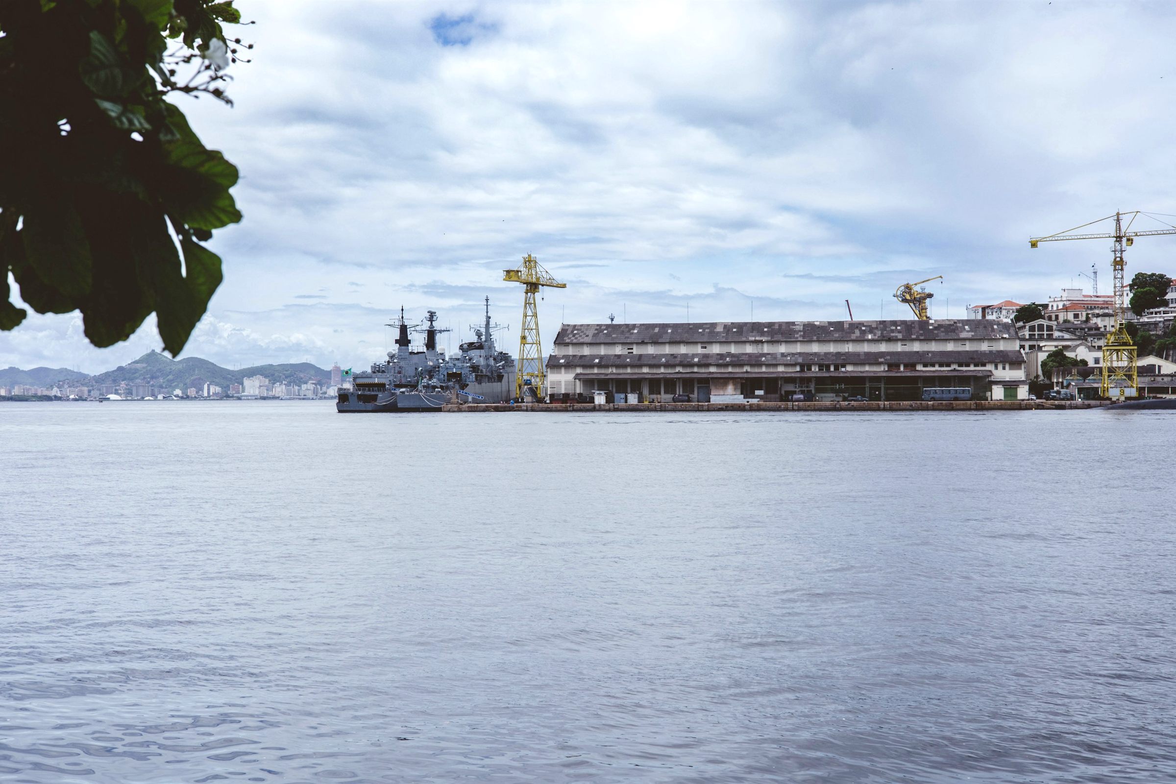 Naval ship docked at a shipyard with cranes across the water